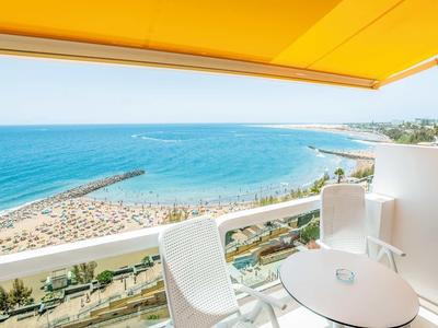 Balkon mit weißem Tisch und Stühlen, gelbes Dach, Blick aufs blaue Meer und Sandstrand.
