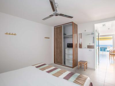 Schlafzimmer mit Doppelbett, gestreifter Tagesdecke, kleinem Spiegel und Blick in einen Flur mit Balkon.