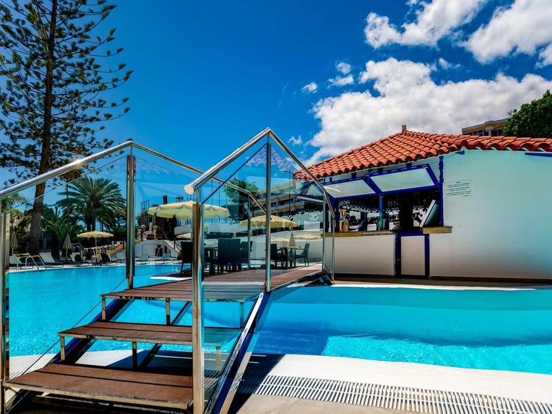 Piscina ricreativa con acqua blu limpida, passerella in legno e terrazza solarium sotto un cielo azzurro.