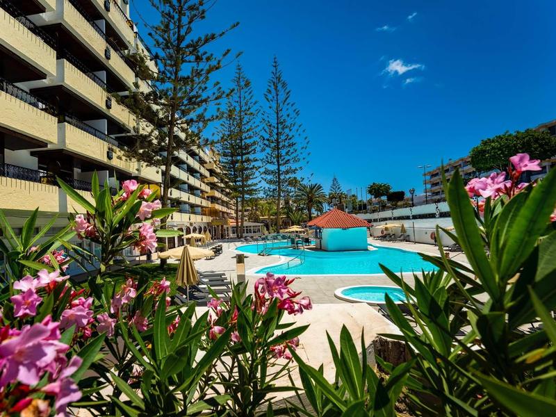 Piscina dell'hotel con fiori rosa circostanti e edificio su più piani sotto un cielo azzurro.
