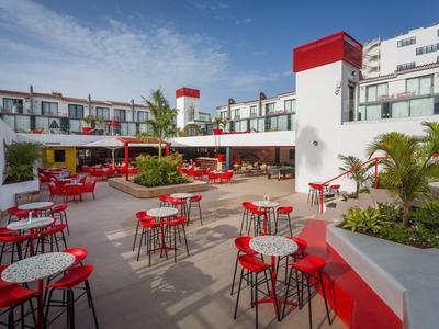 Cortile moderno di un hotel con sedie rosse, tavoli e piante verdi sotto un cielo blu.