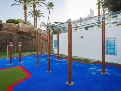 Outdoor-Fitnessbereich mit Stangen und blauer Gummibodenfläche neben Felsen und Palmen.