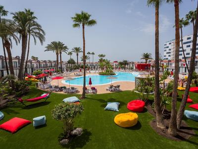 Hotelgarten mit Pool, umgeben von Palmen, Liegestühlen und bunten Sitzkissen auf grünem Rasen.