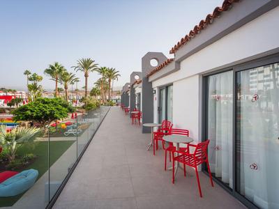 Balkon mit roten Stühlen und Tischen vor Hotelzimmern mit Blick auf Palmen und Pool.