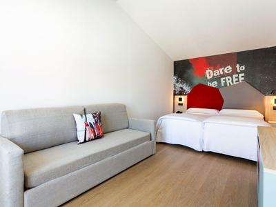 Camera d'hotel luminosa con due letti singoli, divano grigio e decorazione murale con testo.