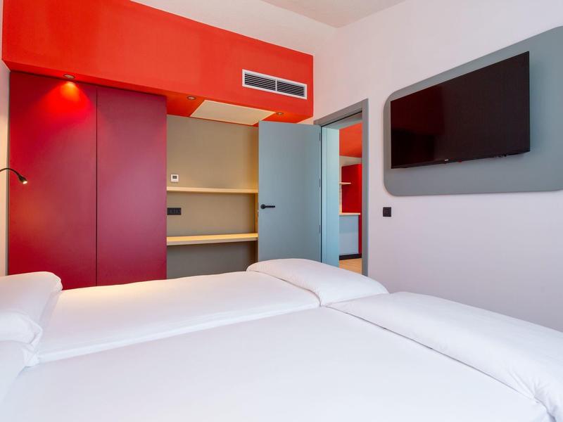 Camera d'hotel moderna con letto matrimoniale, TV a parete e armadio aperto in tonalità rosse e blu.