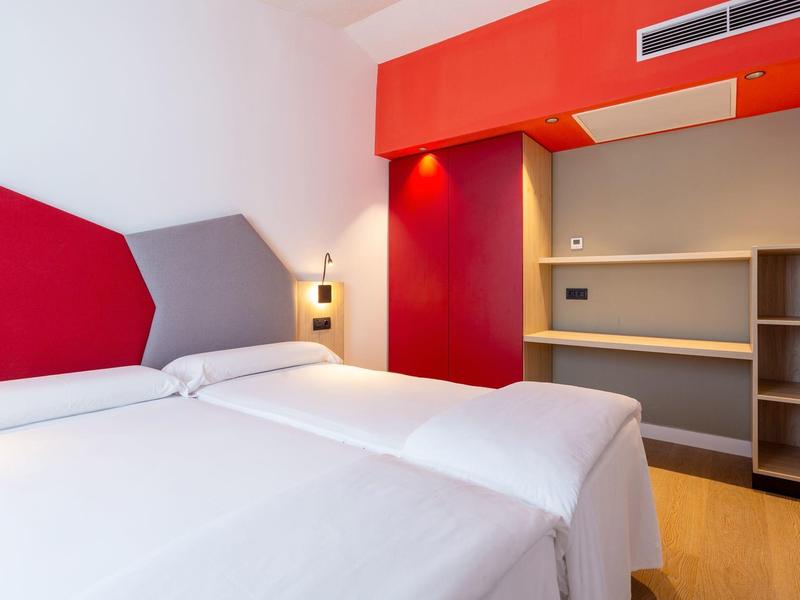 Camera d'hotel moderna con due letti singoli, testata coordinata e scaffalature aperte.