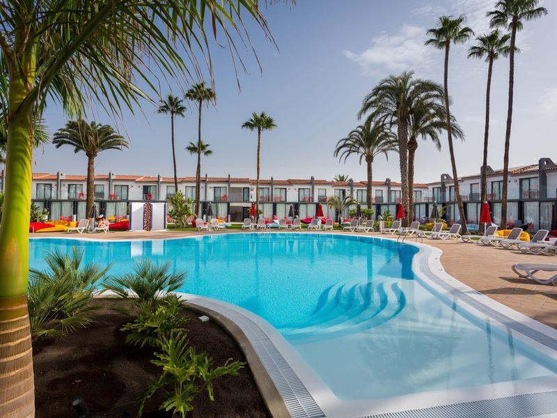 Piscina all'aperto moderna con lettini e palme in un contesto alberghiero tropicale sotto un cielo azzurro.