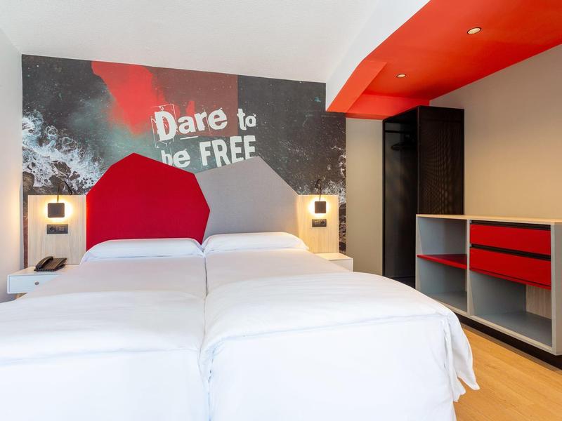 Camera d'hotel luminosa con due letti singoli e accenti rossi su parete e mobili.