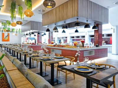 Modernes Restaurantinterieur mit Holztischen, Polstersitzen und grünen Pflanzen an der Decke.