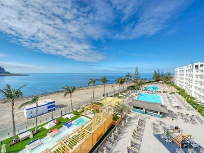 Hotel z basenem i widokiem na plażę w słoneczny dzień z błękitnym niebem i chmurami.