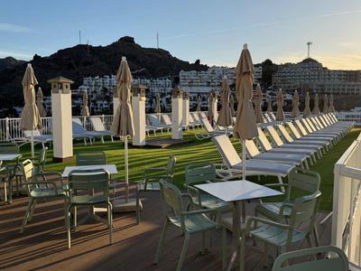 Słoneczny taras z stołami, krzesłami i zamkniętymi parasolami, widok na wzgórza w tle.