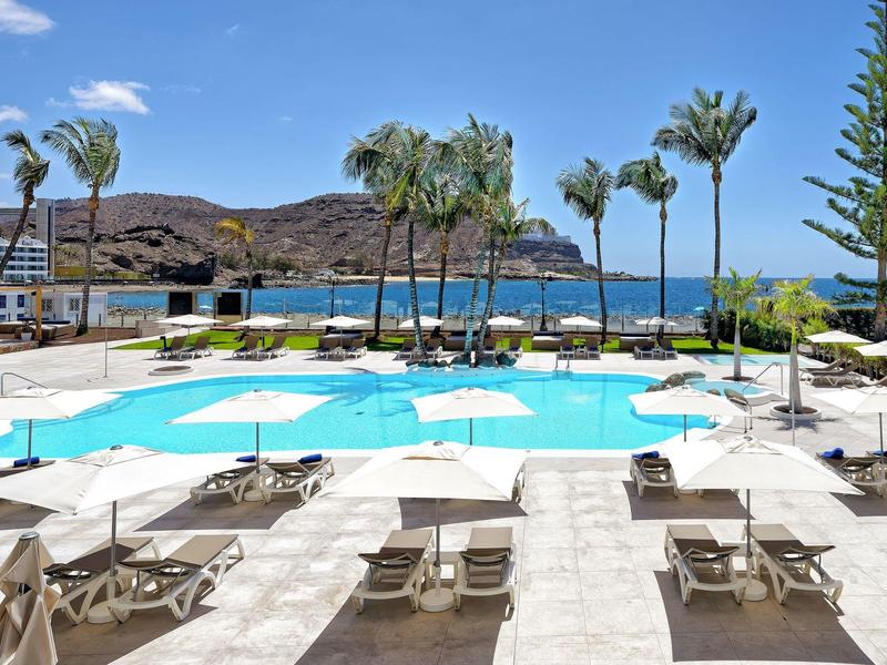 Basen hotelowy z leżakami i palmami z widokiem na morze i góry w tle.