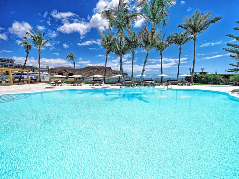 Duży basen z czystą wodą, otoczony palmami i leżakami pod niebieskim niebem.