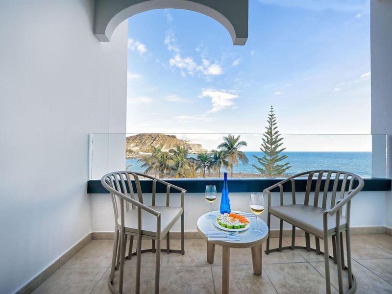 Balkon z dwoma krzesłami i stołem z widokiem na morze, palmy i wzgórza pod czystym niebem.