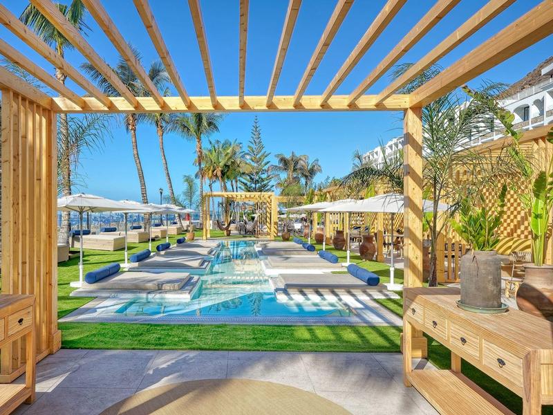 Nowoczesny hotelowy basen z leżakami i drewnianą pergolą, otoczony zielenią i niebieskim niebem.