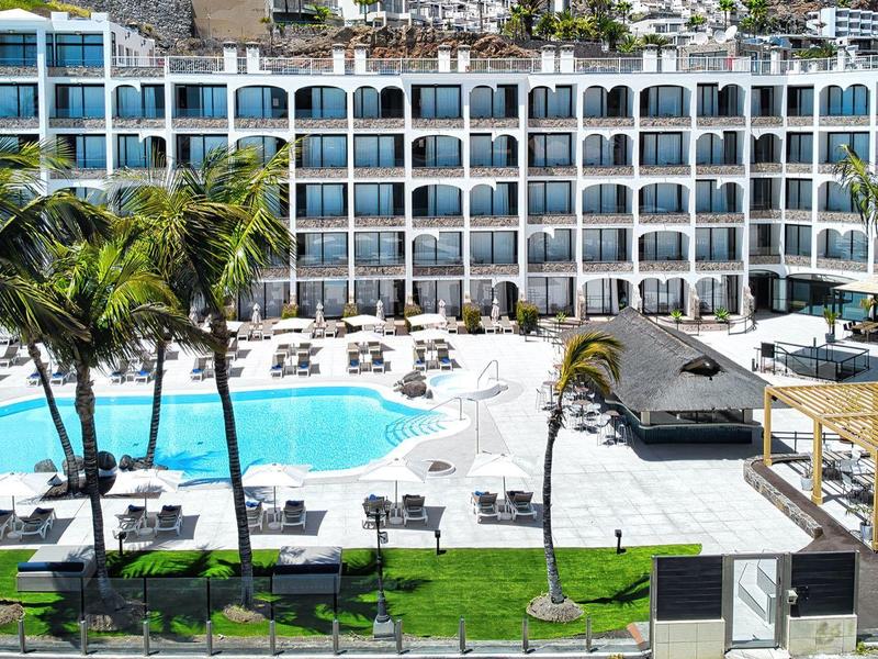 Hotel z dużym basenem, palmami i leżakami przed wielopiętrowym budynkiem z licznymi balkonami.