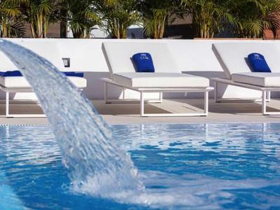 A modern pool with a waterfall and lounge chairs with blue cushions under palm trees.