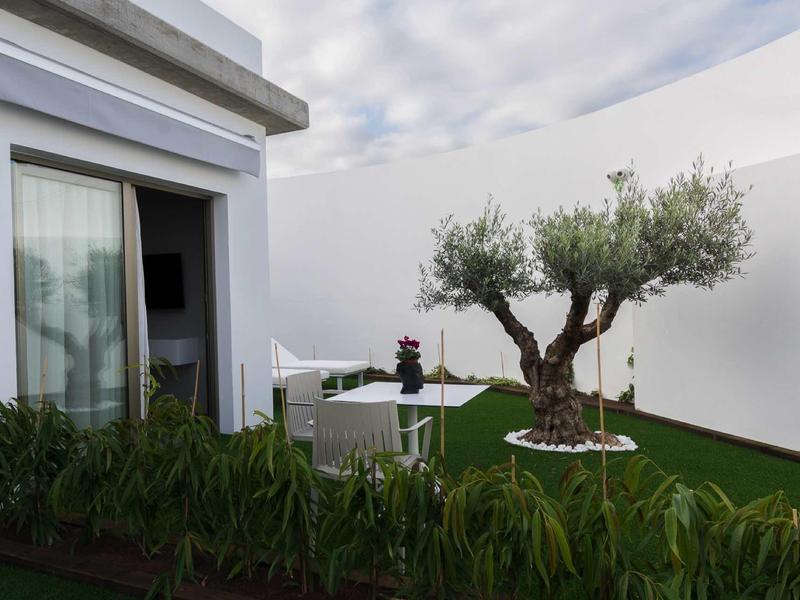 Modern patio with artificial grass, olive tree, and garden furniture against white walls.