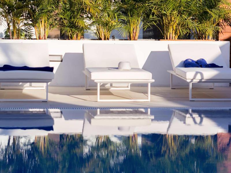 Modern white lounge furniture beside a pool with palm trees in the background.