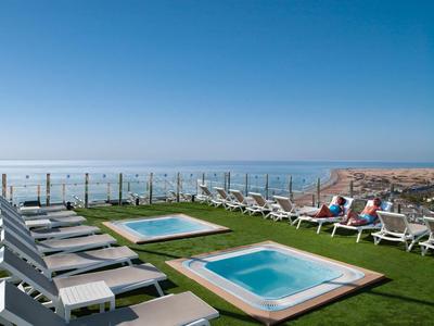 Blick auf zwei kleine Pools mit Liegestühlen auf einer Dachterrasse, im Hintergrund Meer und blauer Himmel.