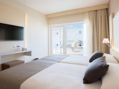 Hell eingerichtetes Schlafzimmer mit großem Bett, braunen Kissen, TV und Balkon mit Meerblick.