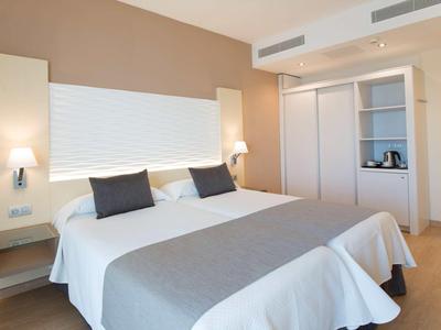 Modernes Schlafzimmer mit großem Doppelbett, weißen Wänden, grauem Bettüberwurf und Nachttischlampen.