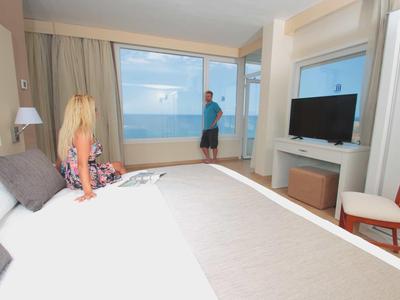 Modernes Hotelzimmer mit großem Bett, Meerblick und zwei Personen, eine Frau sitzt, ein Mann steht.