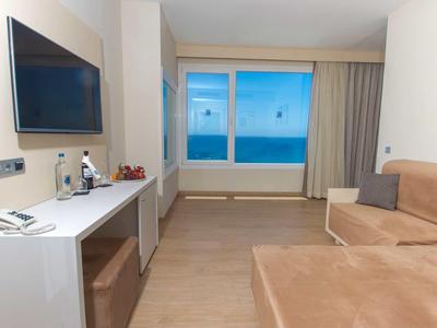Modern eingerichtetes Hotelzimmer mit Fernseher, Schreibtisch, Sofa und großer Fensterfront mit Meerblick.