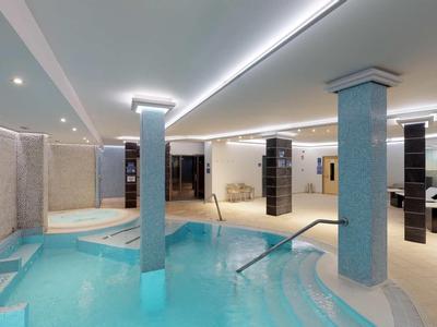 Innenbereich eines modernen Spa mit blauer Poolbeleuchtung, Säulen und Liegestühlen in hellem Raum.
