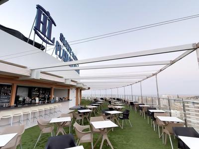 Dachterrasse mit weißen Tischen, schwarzen Stühlen, grüner Rasenmatte und großer Pergola.