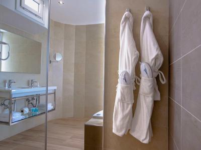 Helles Badezimmer mit zwei weißen Bademänteln, Glaswaschbecken und großen, beige gefliesten Wänden.