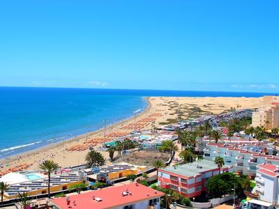 Strand mit goldenem Sand und blauem Meer, umgeben von Gebäuden und grünen Flächen unter klarem Himmel.