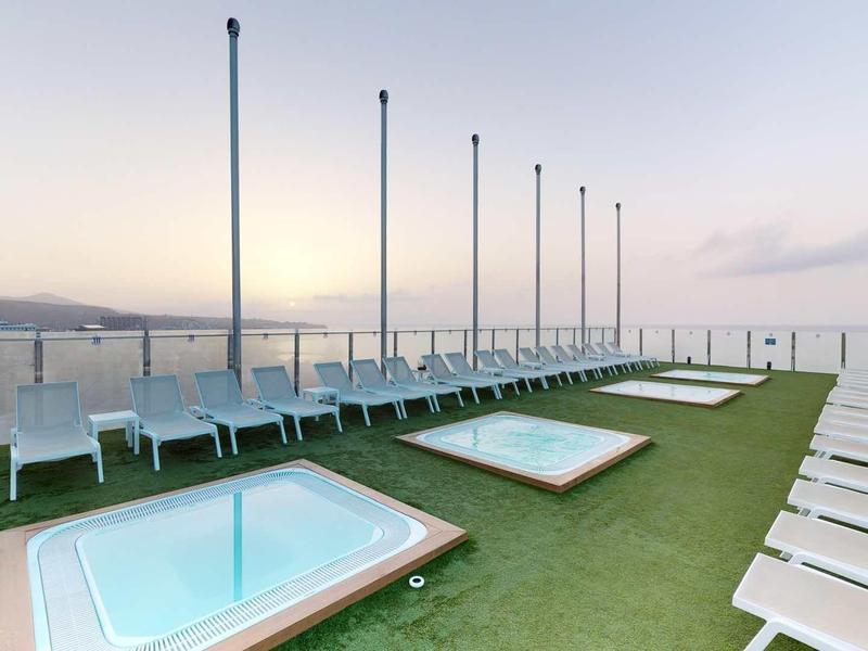 Taras na dachu z dwoma jacuzzi i wieloma leżakami o zmierzchu z widokiem na morze.