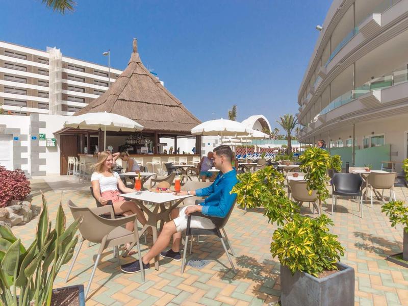 Dwie osoby siedzą na zewnątrz przy stole obok basenu w ośrodku hotelowym w słoneczny dzień.