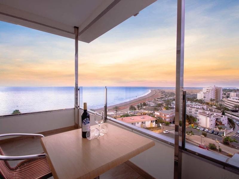 Balkon z stołem, krzesłami i widokiem na plażę, morze i miasto o zachodzie słońca.