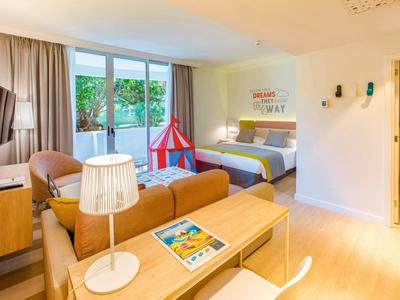 Modernes Hotelzimmer mit Doppelbett, Sofa, Arbeitsplatz, großem Fenster und Kinderzelt, hell und freundlich.