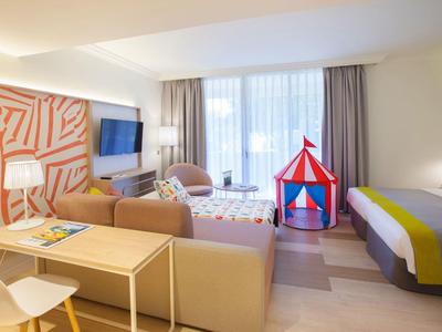 Hell eingerichtetes Wohnzimmer mit Sofa, Tisch, Fernseher, Kinderspielzelt und großem Fenster mit Vorhängen.