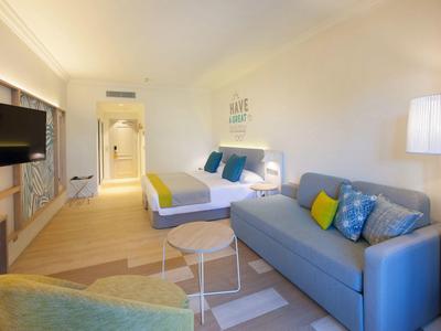 Helles Hotelzimmer mit Doppelbett, blauem Sofa, Fernseher und modernen Möbeln in warmen Farben.