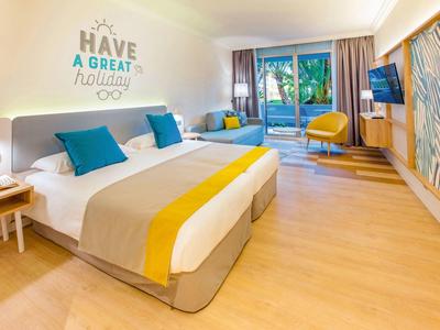 Helles Hotelzimmer mit Doppelbett, blauen Kissen, gelben Akzenten und Zugang zur Terrasse.