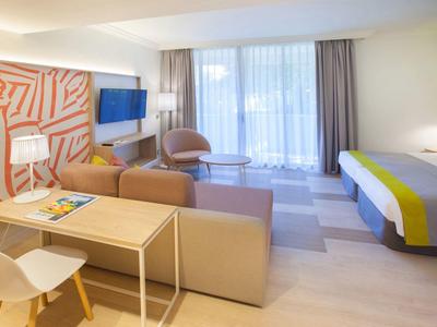 Helles Hotelzimmer mit Doppelbett, Sofa, Schreibtisch, Stuhl und großem Fenster mit Vorhängen.