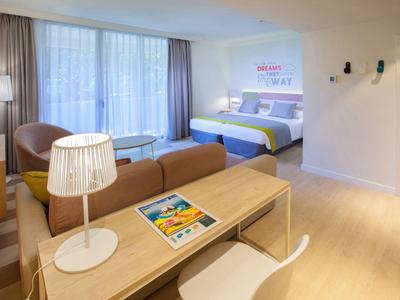 Hell eingerichtetes Zimmer mit Doppelbett, Sofa, Schreibtisch und großen Fenstern mit Vorhängen.