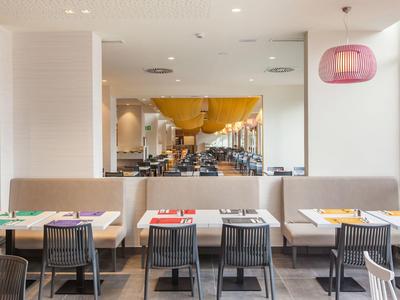 Moderner Gastronomiebereich mit beigen Sitzbänken, schwarzen Stühlen und farbigen Tischsets.
