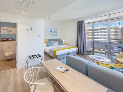 Modernes, helles Hotelzimmer mit Doppelbett, Sofa, Schreibtisch und Balkon mit Stadtblick.