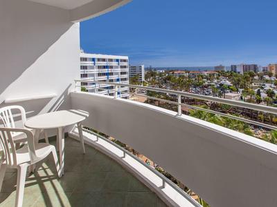 Balkon mit weißem Geländer, zwei Stühlen und kleinem Tisch, Blick auf Wohngebäude und blauen Himmel.