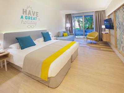 Modernes Hotelzimmer mit Doppelbett, bunten Kissen, Holzfußboden und Glasfenster zur Terrasse.