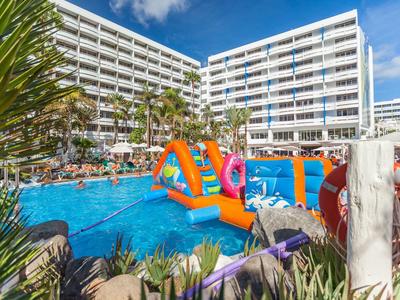 Großes Hotel mit Außenpool, aufblasbarem Wasserspiel und Palmen unter blauem Himmel.