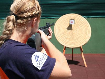 Frau mit blondem Zopf zielt mit einer Pistole auf eine Zielscheibe auf rotem Boden vor grünem Hintergrund.