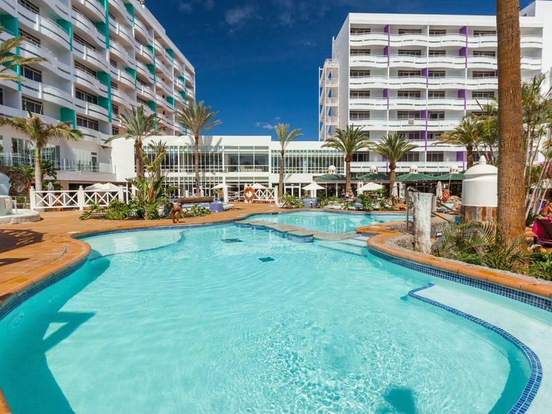 Modern hotel complex with large outdoor pool and sun loungers under blue sky