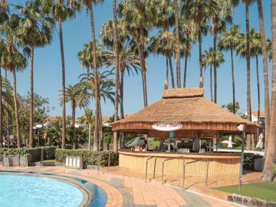 Poolbar mit Strohdach umgeben von Palmen neben blauem Schwimmbecken unter klarem Himmel.